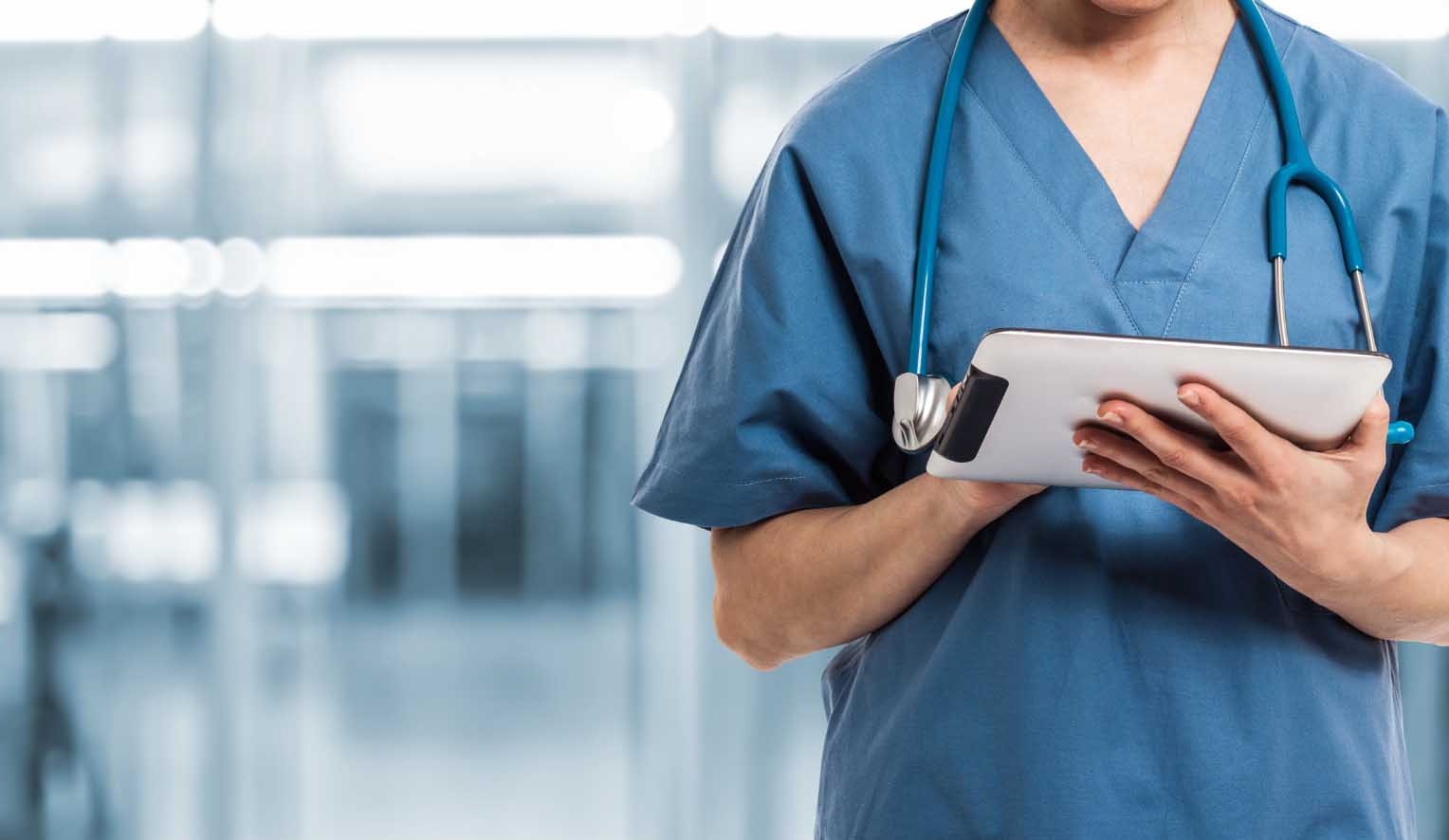Medical professional in scrubs holding a tablet. Stethoscope around their neck matches their blue scrubs. 